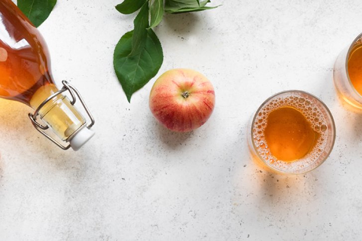 Flasche Apfelsaft, liegend; Gläser gefüllt; umgeben von Äpfeln und Blättern auf hellem Hintergrund.