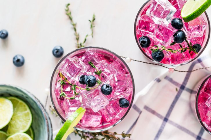 Drei pinke Cocktails mit Limetten und Heidelbeeren stehen auf kariertem Tuch neben Limettenschale.