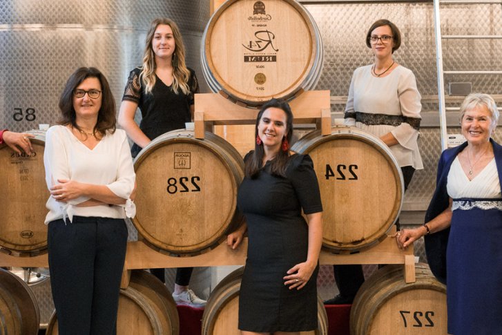 Fässer stehen in einer Reihe; Personen posieren davor in einem Weinkeller mit Metalltanks.