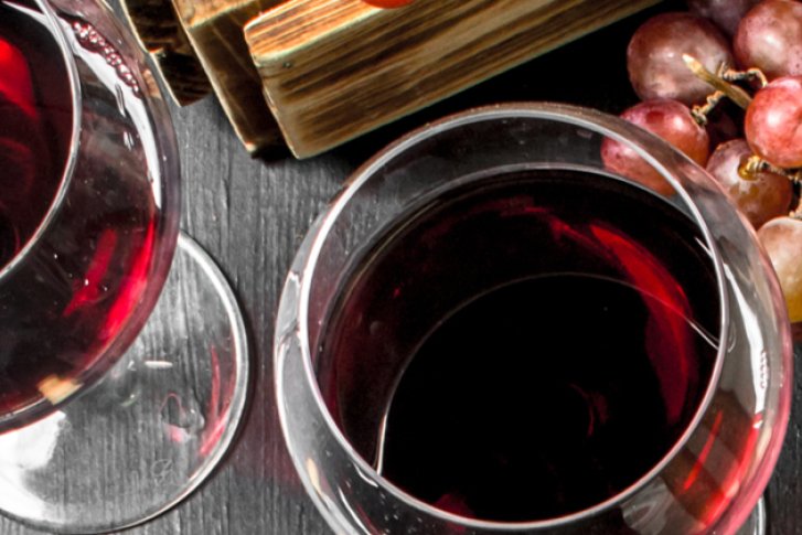 Zwei Weingläser gefüllt mit Rotwein, daneben Weintrauben und ein Flaschenverschluss auf dunklem Holztisch.