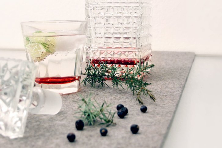 Karaffe mit Glas, auf grauer Unterlage stehende, mit Gin, Wacholder und Limette gefüllt.