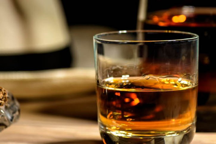 Glas mit Whisky steht auf Tisch, daneben Zigarre und Strohhut im Hintergrund.