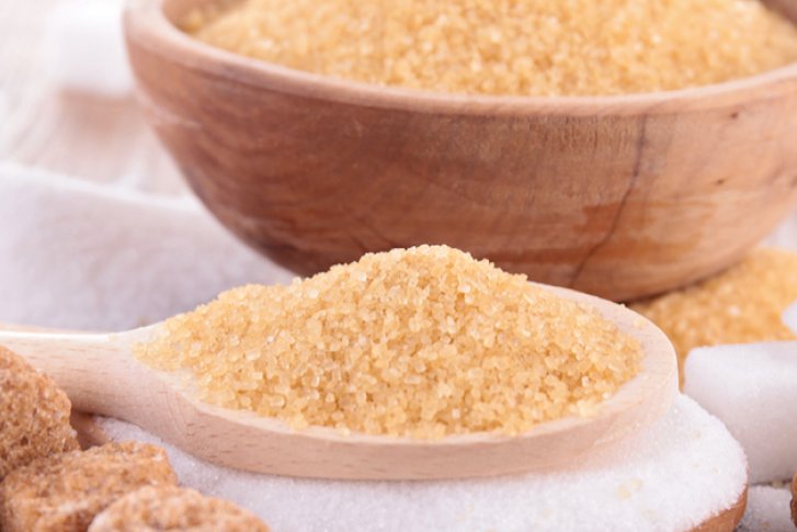 Holzlöffel mit braunem Zucker, umgeben von Würfelzucker, auf Holztisch neben Holzschale.