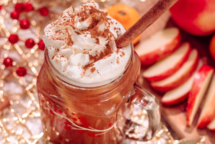 Heißgetränk mit Sahne und Zimt in Glas; daneben Äpfel, Zimtstangen, Dekoration.