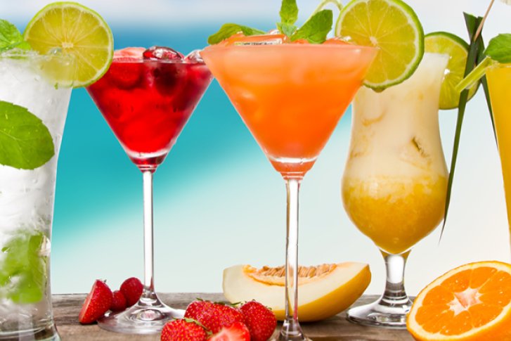 Fünf bunte Cocktails stehen dekorativ auf einem Holztisch vor einem unscharfen Strandhintergrund.