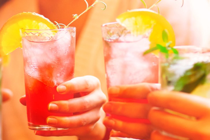 Gläser mit buntem Cocktail und Eis, gehalten von Händen, in geselligem, warmem Ambiente.