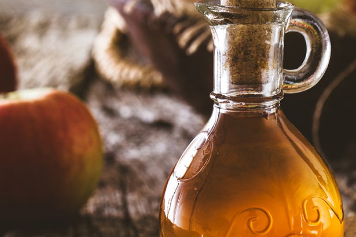 Karaffe mit Apfelsaft steht rustikal auf Holztisch, umgeben von frischen roten Äpfeln.