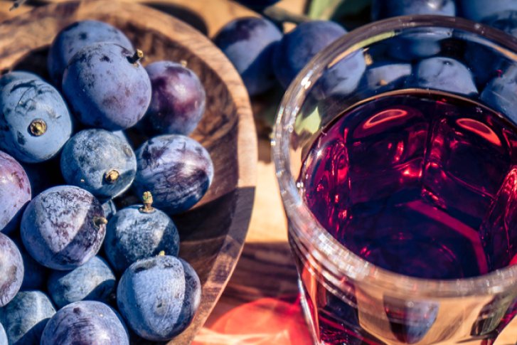 Pflaumen liegen neben einem Glas roten Getränks auf einem Holztisch im Sonnenlicht.