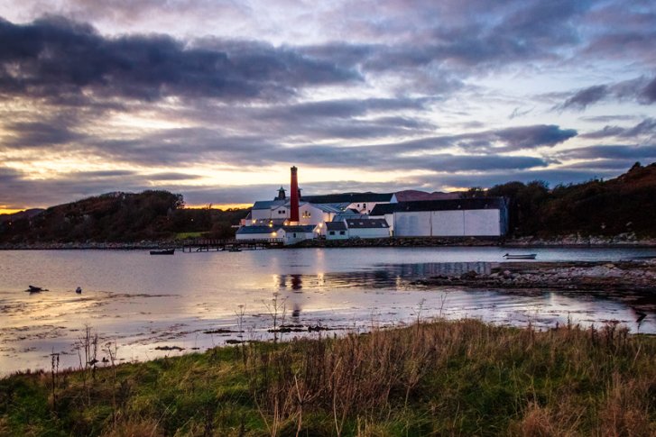 Brennerei am Wasser mit Schornstein, von Gräsern umgeben, im Abendlicht unter bewölktem Himmel.
