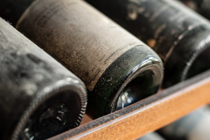 Verstaubte Weinflaschen liegen in einem hölzernen Regal, teilweise unleserliche Etiketten sichtbar.