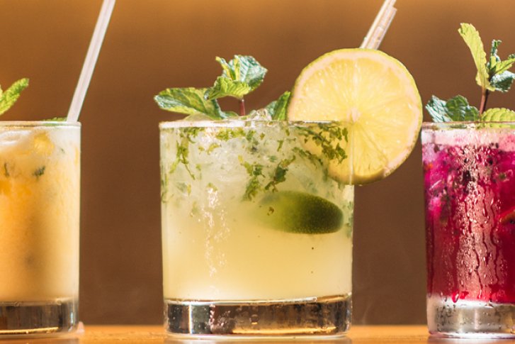 Drei Cocktails mit Obstgarnitur stehen auf einer Holztheke vor einem unscharfen Hintergrund.