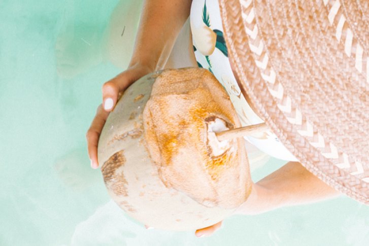 Eine Person hält eine Trinkkokosnuss im türkisfarbenen Wasser, trägt einen großen Strohhut.