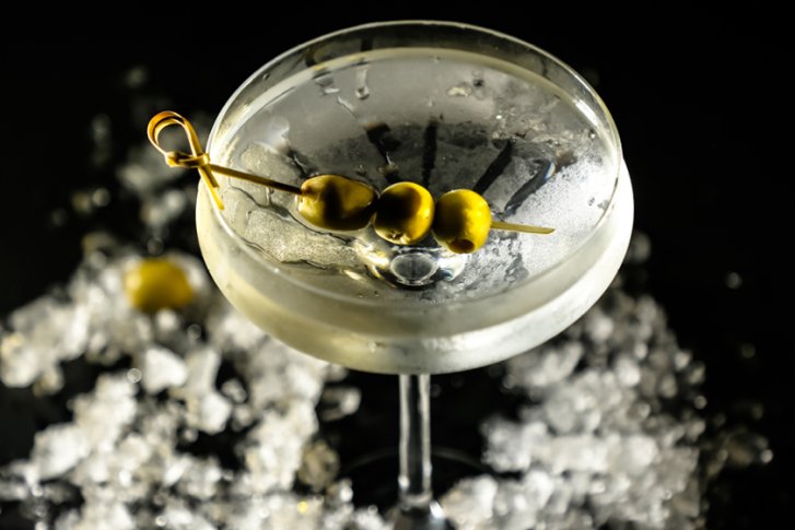 Klassisches Cocktailglas mit Oliven auf einem Spieß, umgeben von zerkleinertem Eis, auf dunklem Hintergrund.