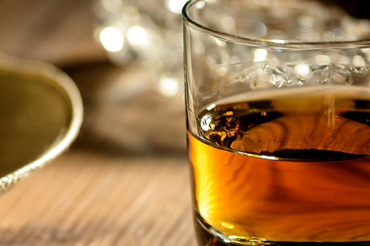 Glas mit brauner Flüssigkeit neben Strohhut auf einem Holztisch, beleuchtet.