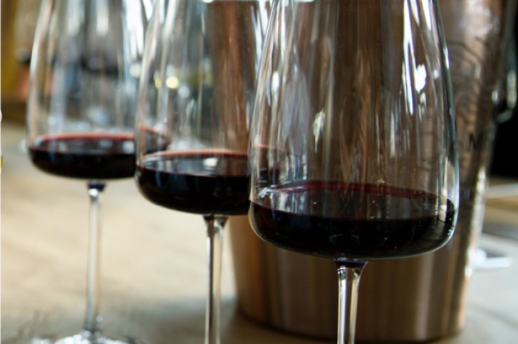 Drei Weingläser mit Rotwein stehen auf einem Holztisch vor einem metallischen Hintergrund.