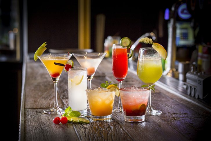 Verschiedene bunte Cocktails stehen dekorativ auf einer Holztheke in einer Barumgebung.