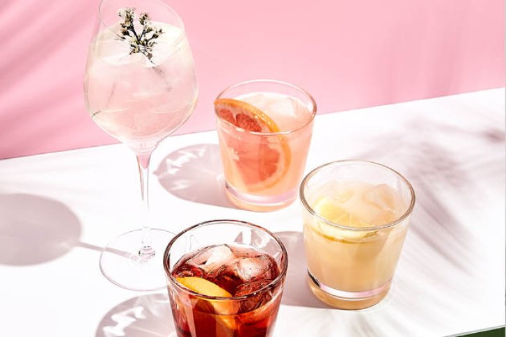 Vier verschiedene Cocktails stehen auf einem weißen Tisch vor einem rosa Hintergrund.
