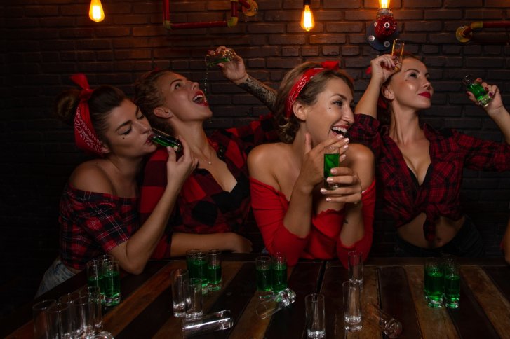 Vier Frauen trinken grüne Shots fröhlich an einem Holztisch in einem gedämpft beleuchteten Raum.