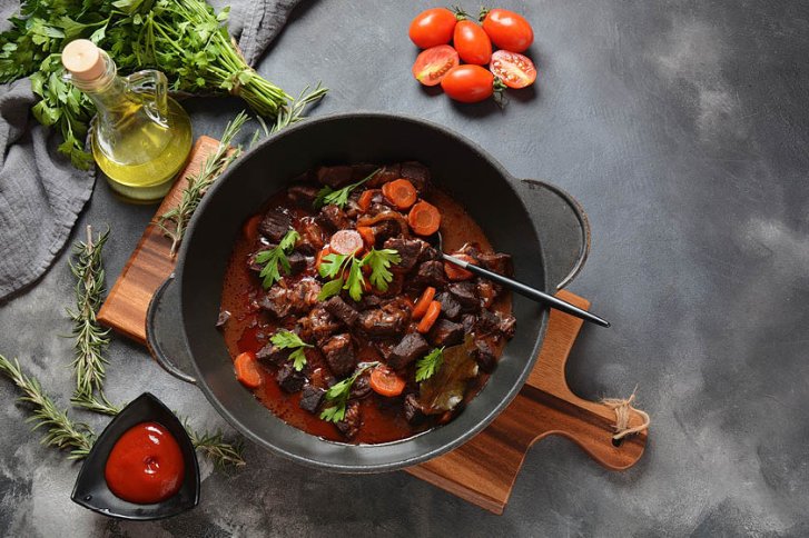 Gusseisentopf mit Fleisch und Karotten, dekoriert mit Petersilie, auf Holzbrett in Küchenumgebung.