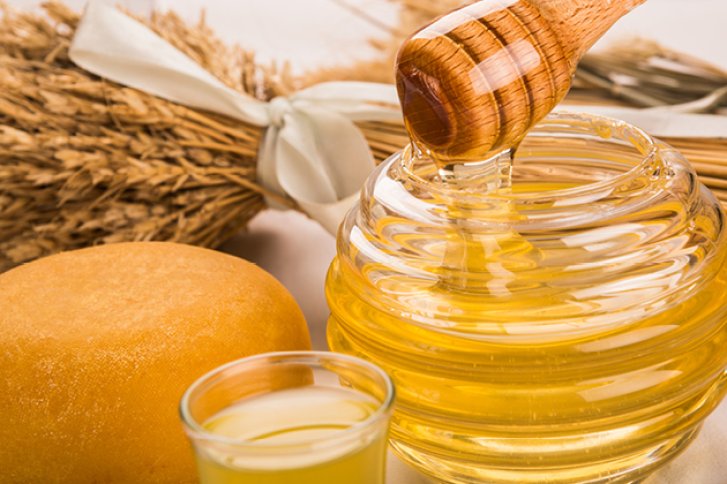 Ein Honigglas wird mit einem Holzlöffel umgerührt, daneben Käse und Weizenbündel.