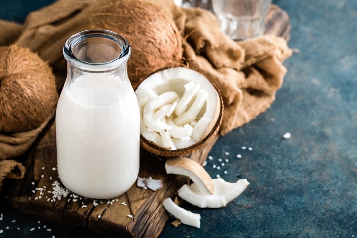 Flasche Kokosmilch steht auf Holzbrett neben geöffneten Kokosnüssen und Tuch auf blauer Oberfläche.