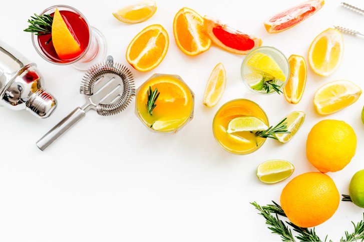 Cocktailgläser stehen mit Zitrusfrüchten und Kräutern dekoriert auf weißem Untergrund, umgeben von Shaker.