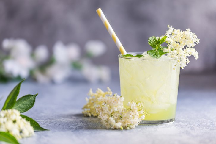 Glas mit gelbem Getränk und Strohhalm, dekoriert mit Blüten, auf grauem Hintergrund.