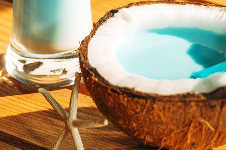 Kokosnusshälfte mit blauem Getränk, auf einem Holzbrett, neben einem hohen Glas.