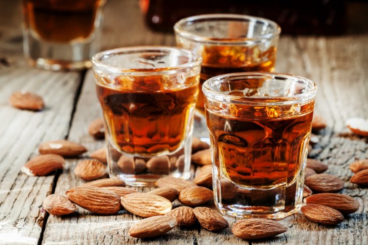 Drei Gläser mit braunem Likör stehen auf Holztisch, umgeben von Mandeln.