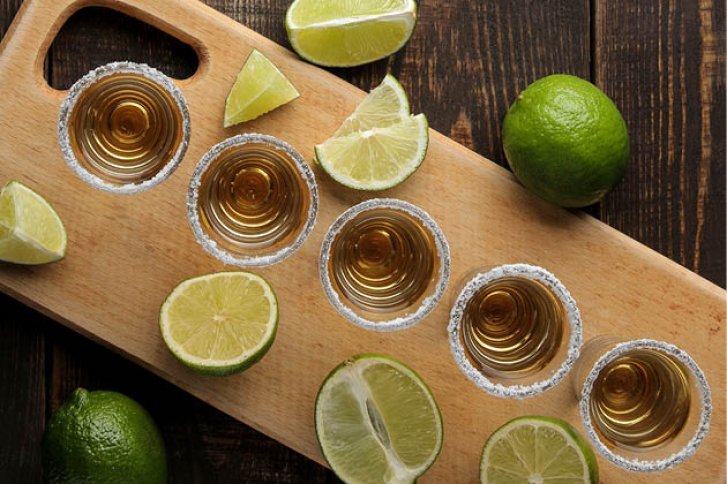 Sechs gefüllte Shotgläser mit Salzrand stehen auf Holzbrett, Limettenscheiben daneben verteilt.