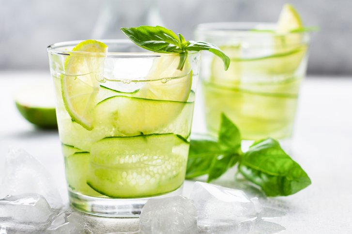 Glas mit Gurken-Limetten-Getränk und Basilikum auf Eiswürfeln, im Hintergrund ein weiteres Glas.