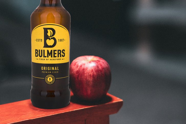 Flasche Bulmers Cider steht neben einem roten Apfel auf einem Holztisch.