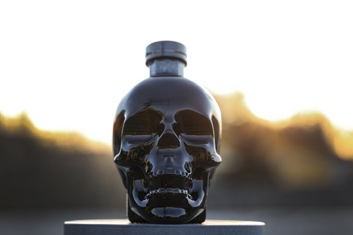 Schwarze, schädelartige Glasflasche steht vor unscharfem Hintergrund, während die Sonne untergeht.