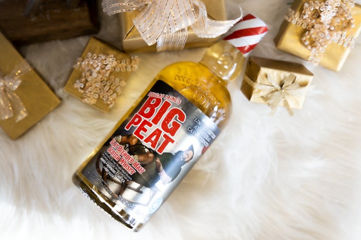 Flasche Whisky liegt auf weißem Fell, umgeben von goldenen Geschenkboxen mit Schleifen.