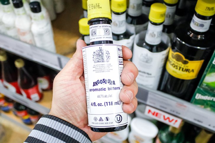 Eine Hand hält eine Angostura Bitterflasche vor einem Supermarktregal mit weiteren Flaschen.