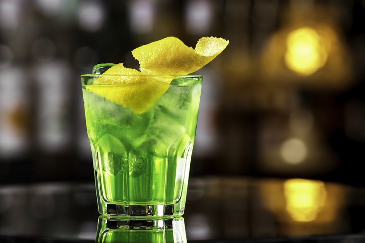 Grünes Getränk mit Eis und Zitronenschale in einem Glas, auf einem reflektierenden Tisch.