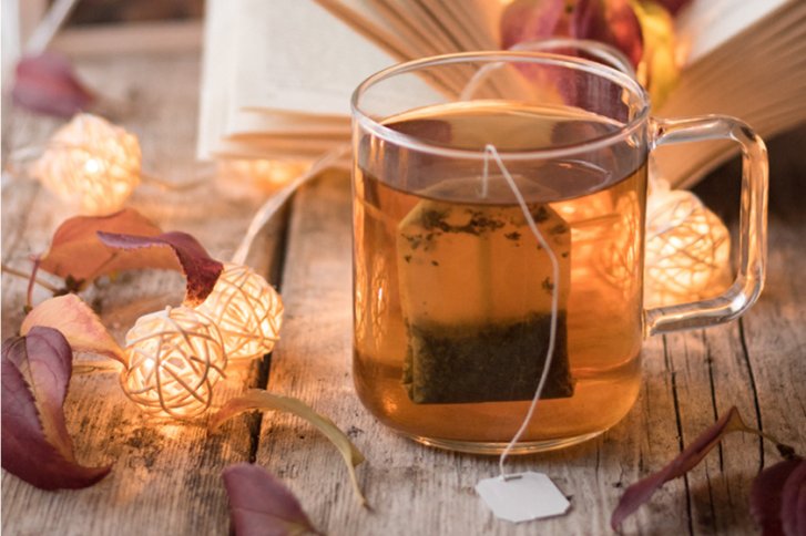 Teetasse steht auf Tisch, umgeben von Herbstblättern und Lichterkette. Buch ist geöffnet.