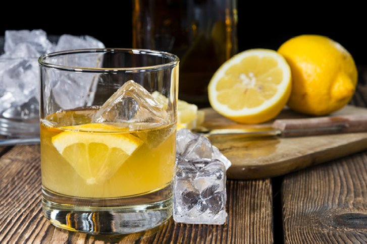 Ein Glas Whisky mit Eis und Zitrone steht auf rustikalem Holztisch neben Zitronenscheiben.