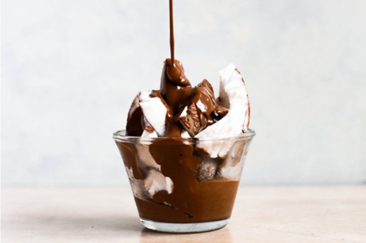 Ein Dessertglas gefüllt mit Schokolade und Kokosnuss, Schokolade fließt herunter, vor hellem Hintergrund.