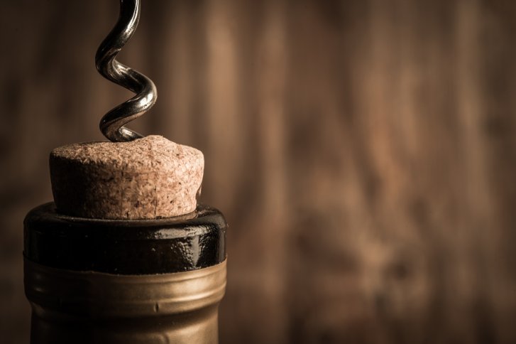 Korkenzieher durchsticht Korken einer Weinflasche vor unscharfem, braunem Hintergrund.