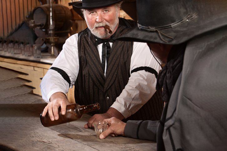 Mann gießt Getränk in Glas, Barumgebung mit rustikalem Charme, trägt Hut und Weste.