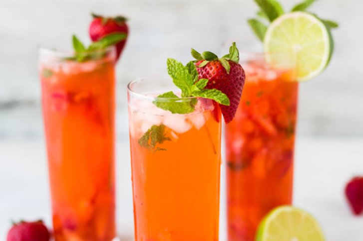 Drei hohe Gläser mit rotem Getränk, garniert mit Erdbeeren und Minze, auf hellem Tisch.