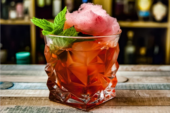 Ein Kristallglas enthält roten Cocktail mit Minze und Zuckerwatte, auf einem Holztisch stehend.