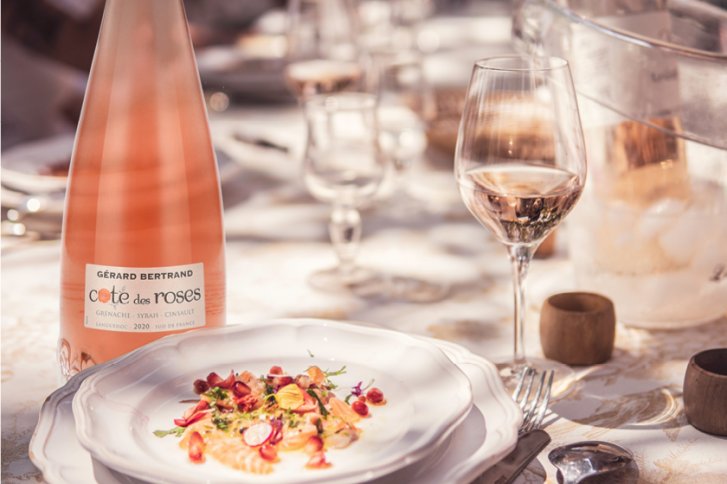 Eine Flasche Roséwein steht neben einem gefüllten Weinglas, umgeben von Gedeck auf Tisch.