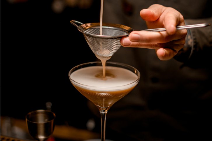 Cocktail wird durch Sieb in Glas gefiltert, auf dunklem Hintergrund mit Hand.