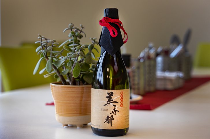 Flasche Sake steht neben Pflanze auf einem Tisch mit rotem Tischläufer im Hintergrund.