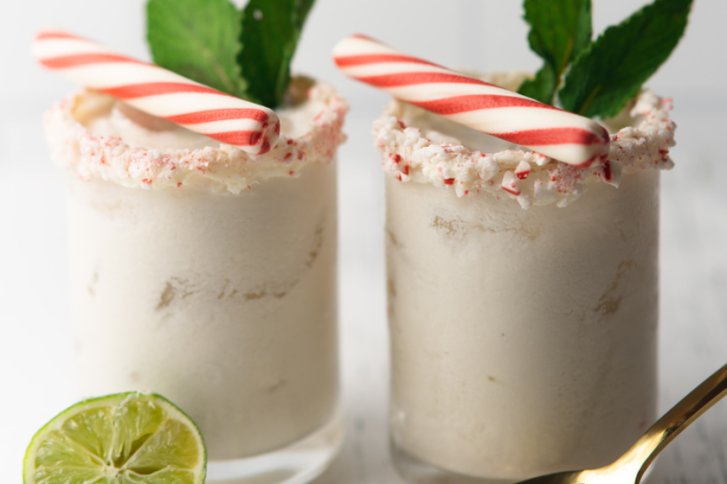 Zwei weiße Getränke in Gläsern mit Pfefferminzstangen, Sahnerand und Minzblättern auf weißem Tisch.