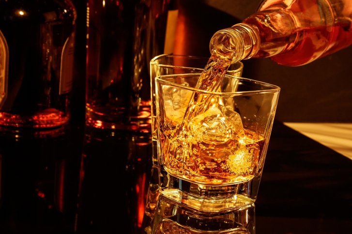 Whiskey wird in ein Glas mit Eiswürfeln gegossen, umgeben von Flaschen, auf dunklem Hintergrund.