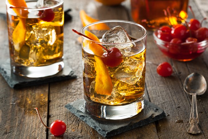 Zwei Whiskeygläser mit Eis und Kirschen stehen auf Schiefertafeln auf einem rustikalen Holztisch.