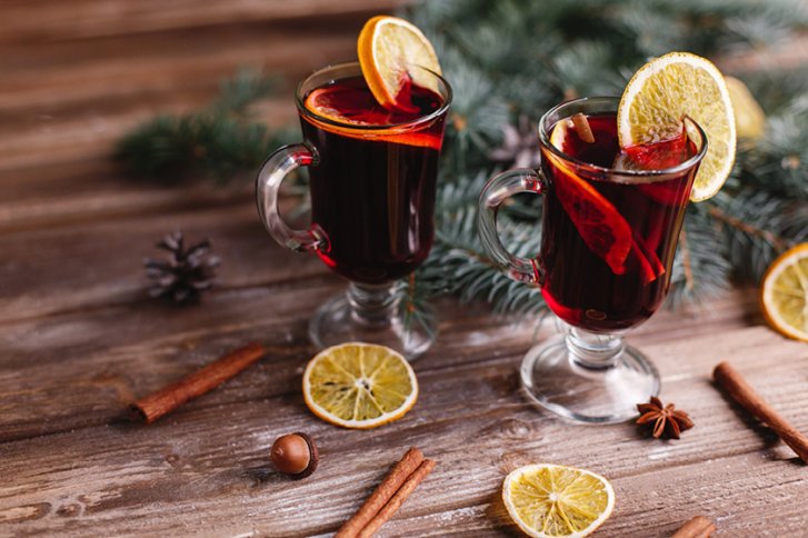 Zwei Gläser Glühwein mit Orangenscheiben stehen auf Holztisch, umgeben von Tannenzweigen und Gewürzen.